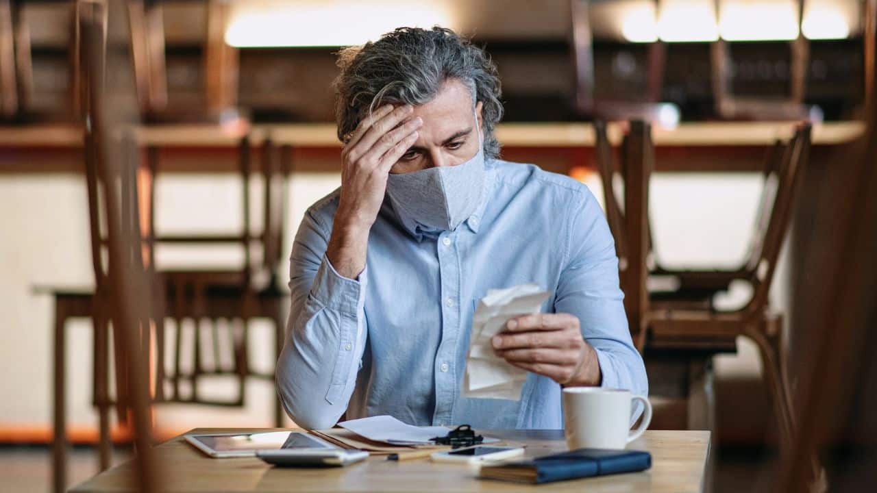 A worried man in a mask looks at receipts, showing financial stress.