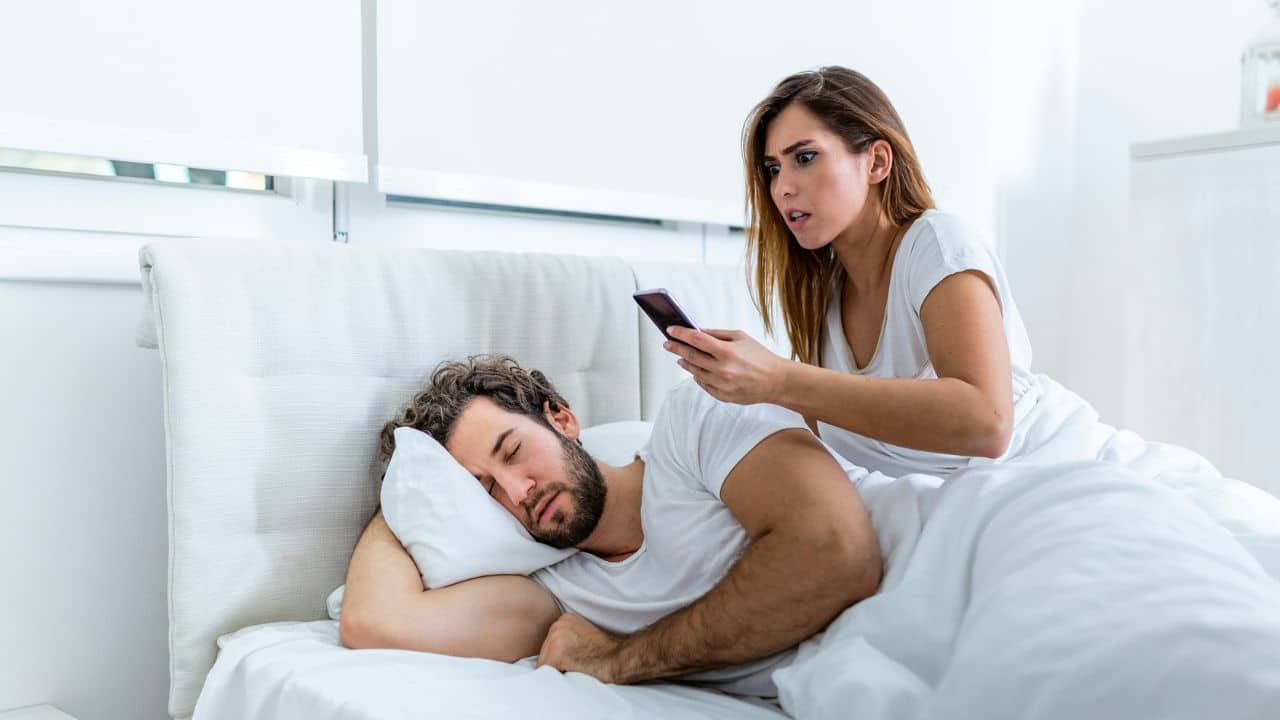 A woman secretly checks her sleeping partner's phone, showing trust issues.