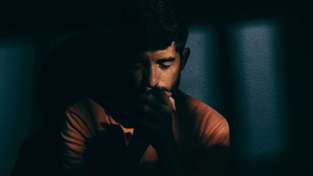 A man sits alone in the dark, with his head bowed and hands clasped.
