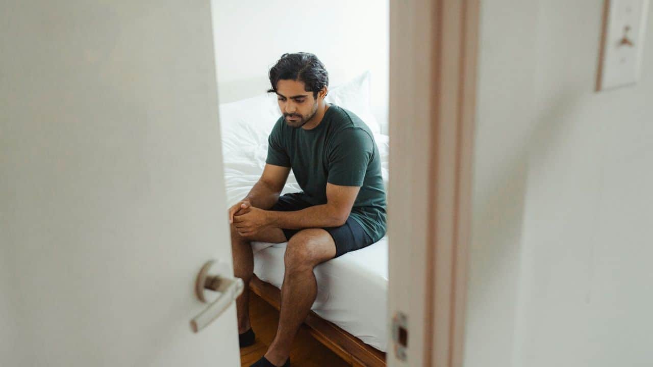 A man sits on the edge of a bed, looking down with his hands clasped together.