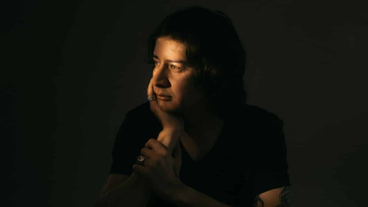 A person with dark hair looks to the side, resting their chin on their hand.