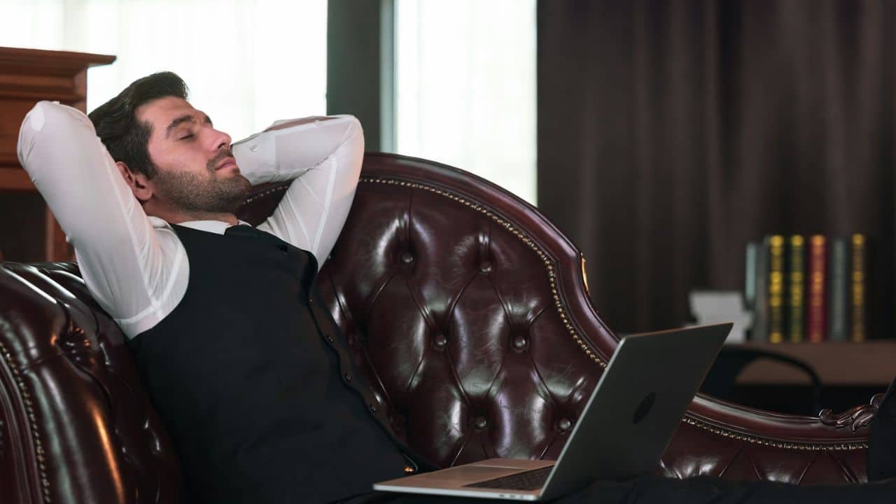 A man in a vest and white shirt reclines on a leather couch with his hands behind his head.