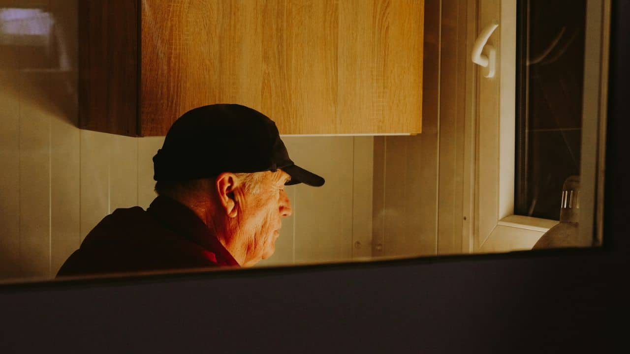 A side profile of an older man in a baseball cap looks out a window.