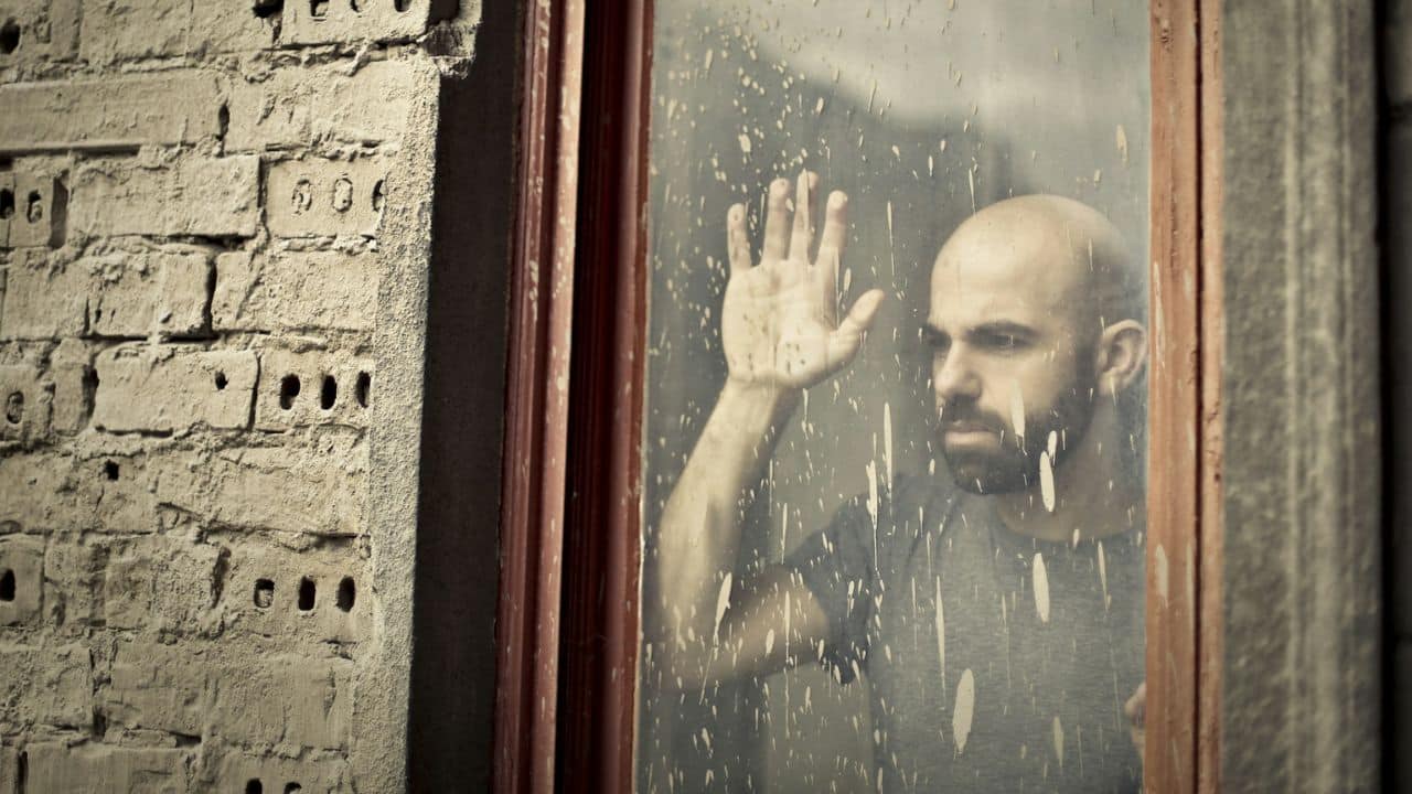 A bald man with a beard looks out a dirty window with his hand on the glass.
