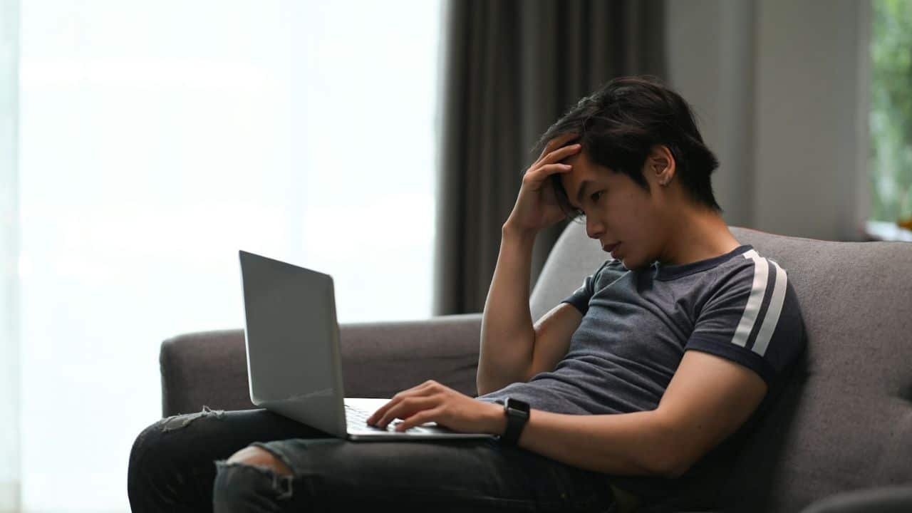A distressed man sits on a couch with a laptop, his hand on his head.