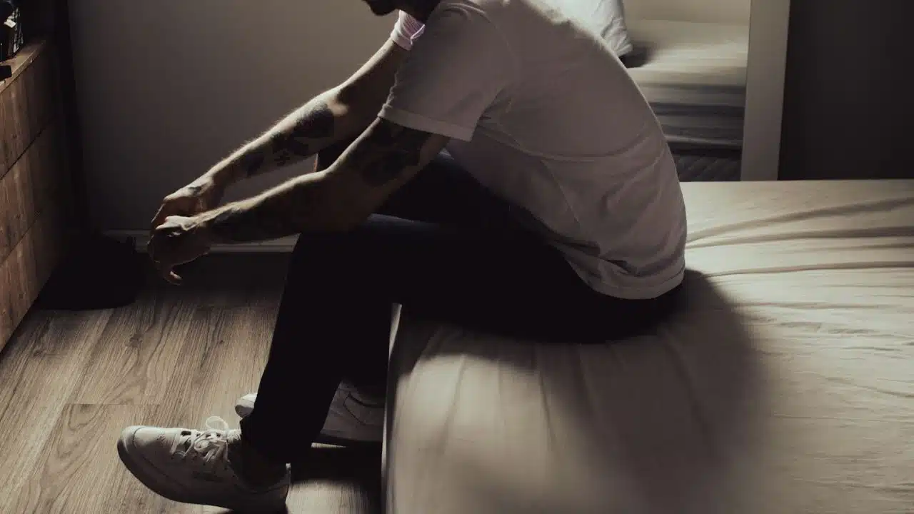 A man with tattooed arms sits on the edge of a bed, looking down.