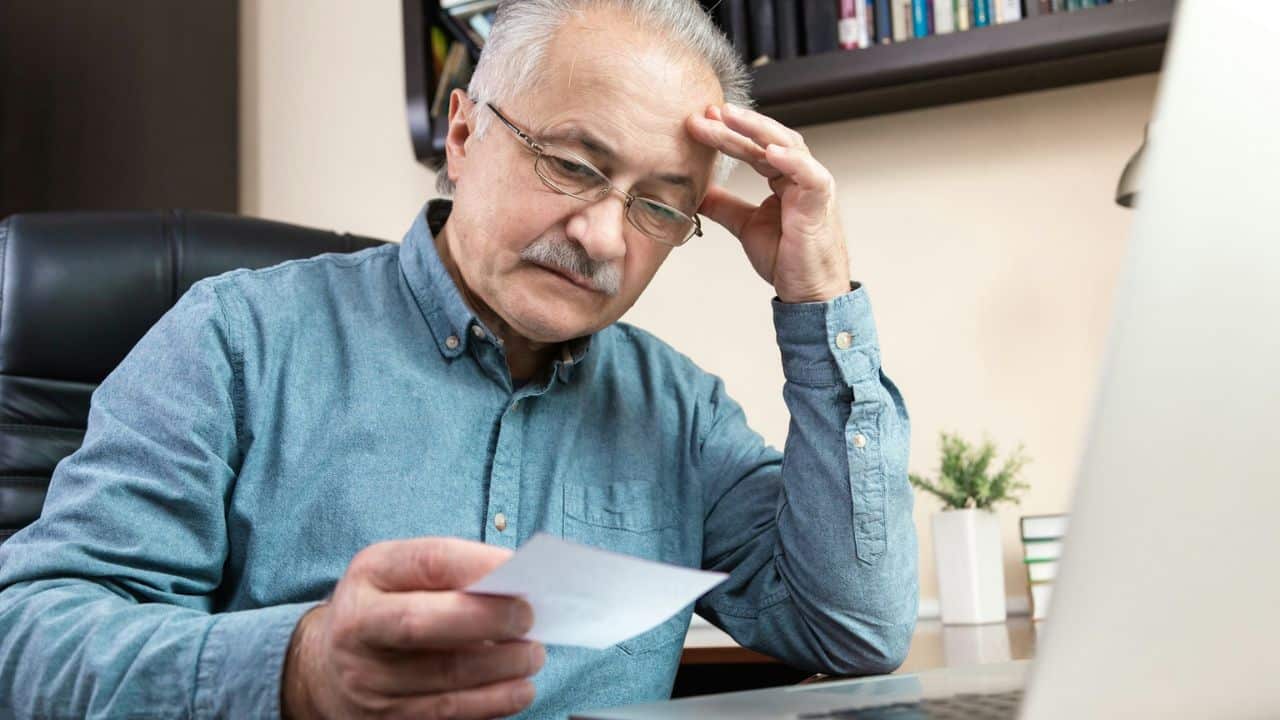 An older man in a blue shirt with his hand on his head looks at a piece of paper.