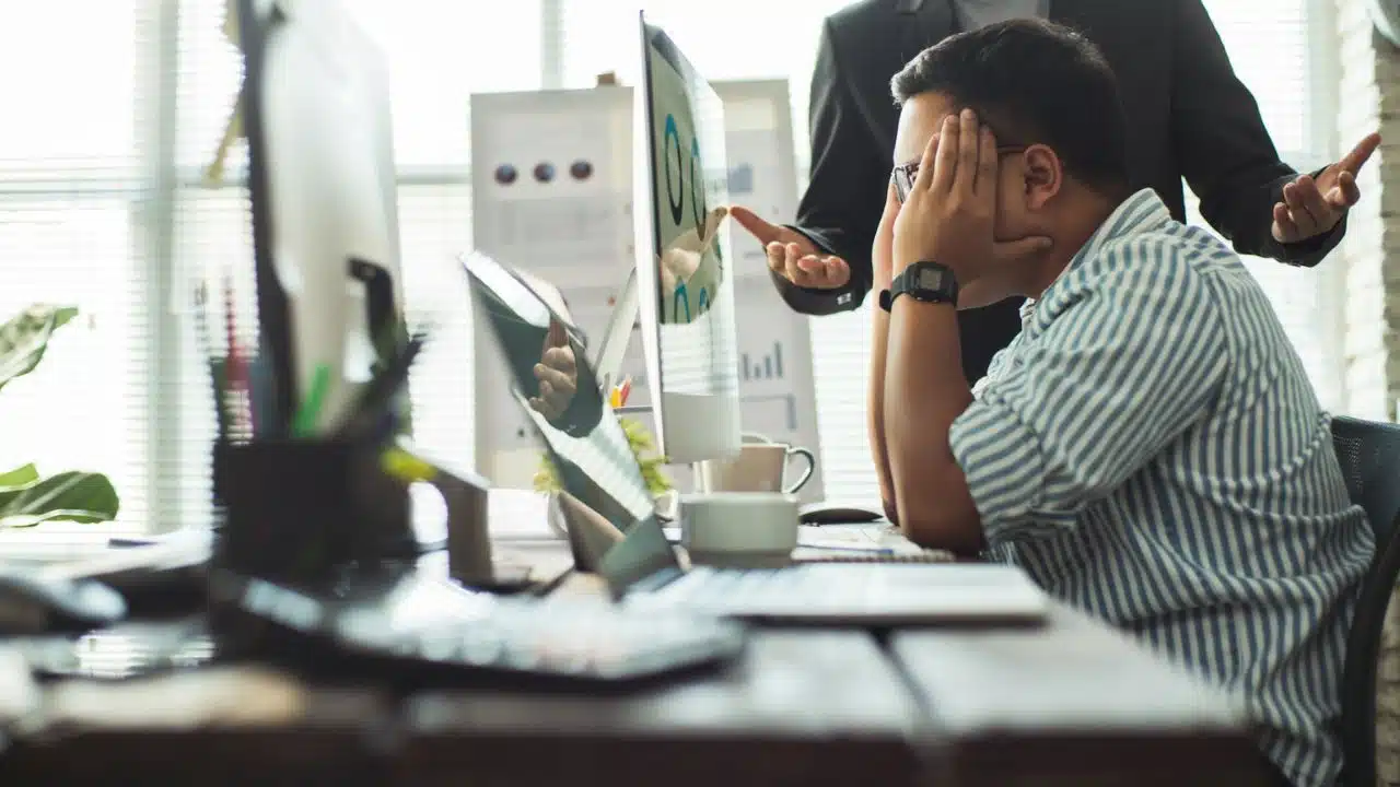 A stressed man with glasses holds his head while a person in a suit points at a computer monitor.