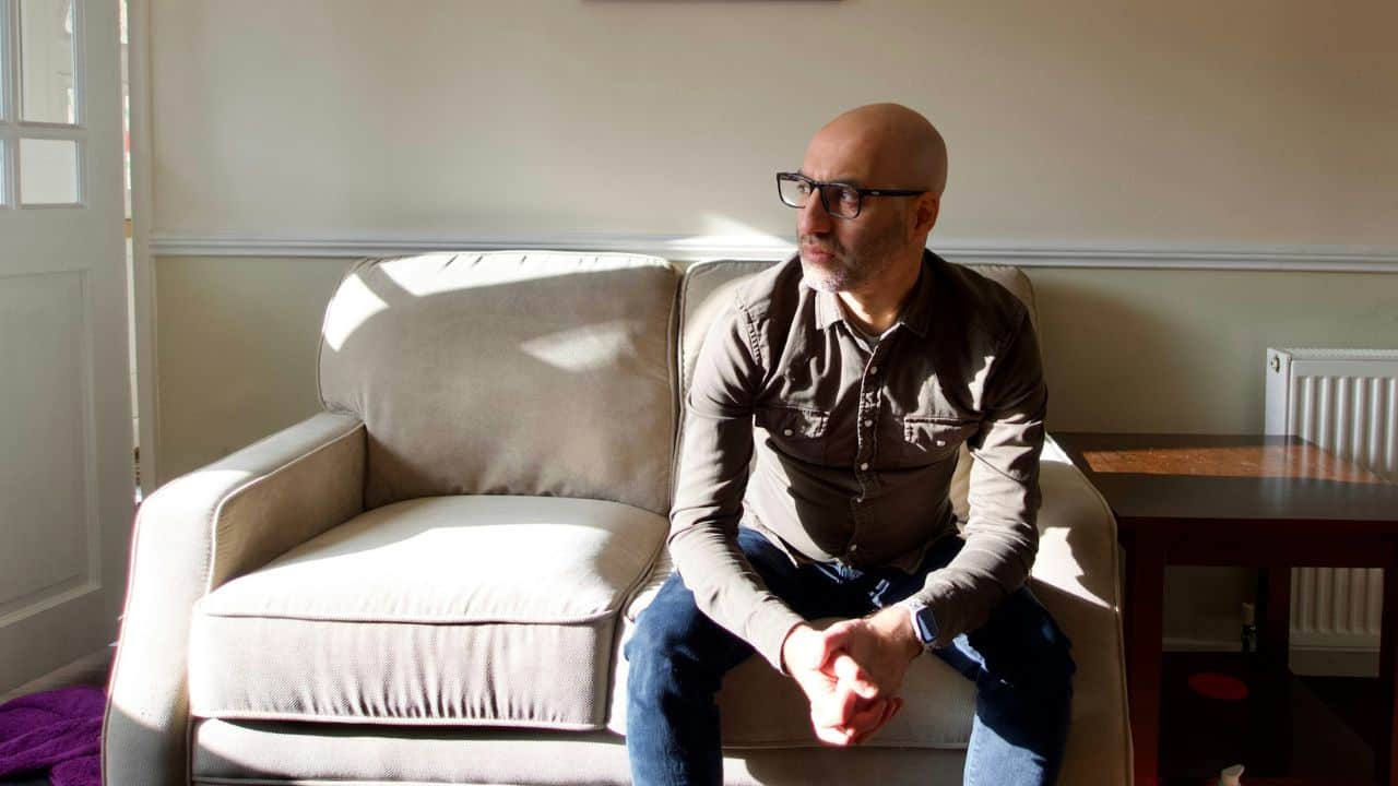 A pensive, bald man with glasses sits on a couch in a sunlit room.