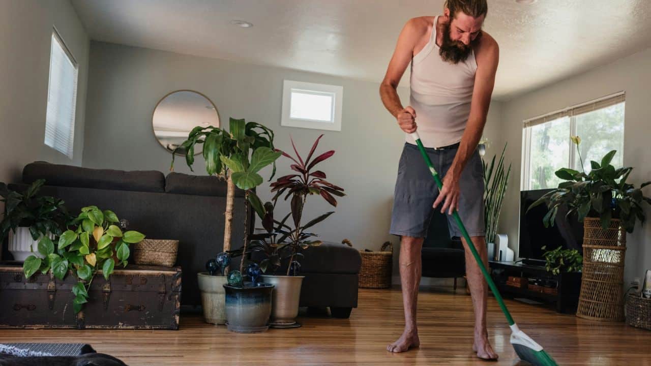 A long-haired man with a beard is sweeping a hardwood floor in his living room.