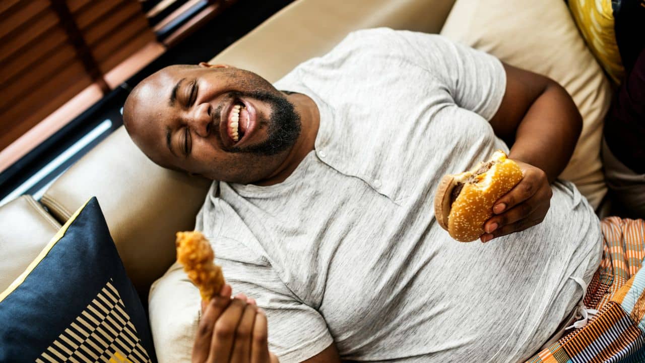 A laughing person is lying on a couch, holding a burger and a piece of fried chicken.