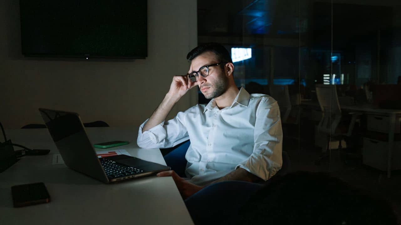 A serious man with glasses is sitting at a desk and working late at night.