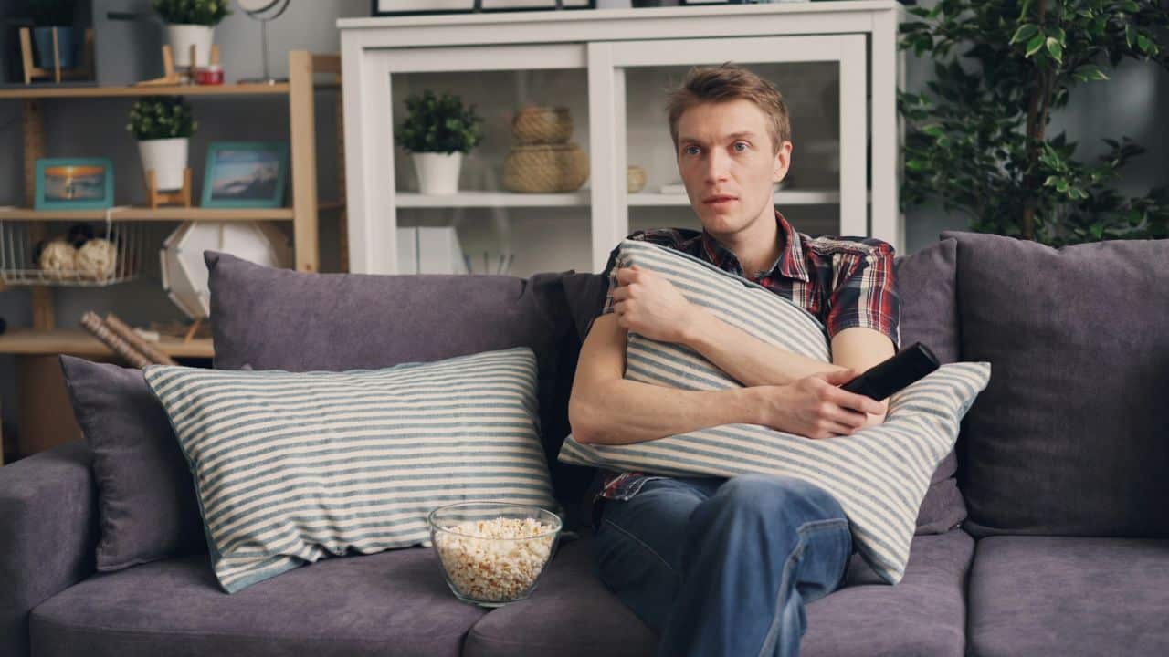 A blank-faced man holding a pillow and a remote sits on a couch.