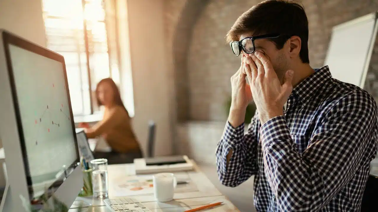 A stressed man in glasses is rubbing his eyes at his desk.