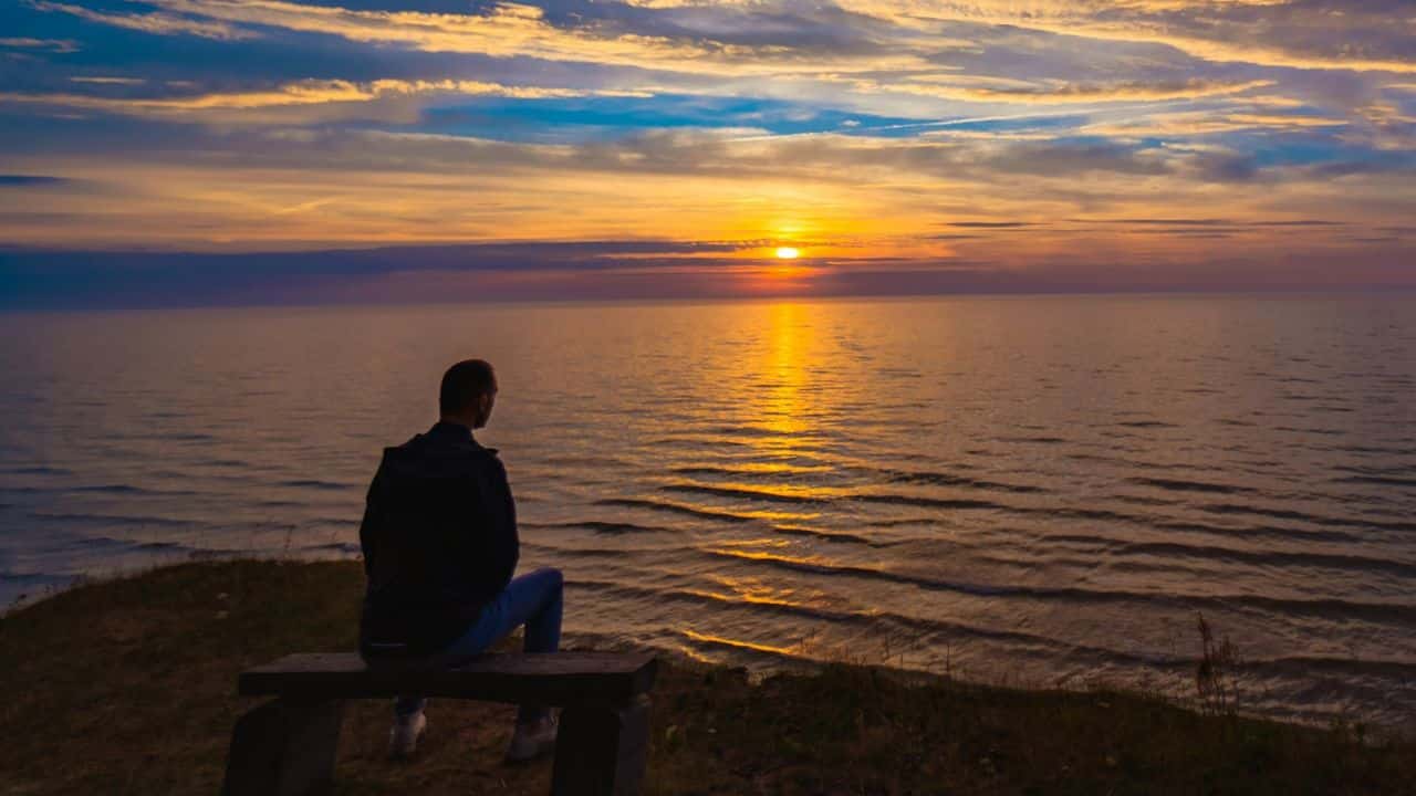 A man sits alone on a bench, looking at a beautiful sunset over the sea.