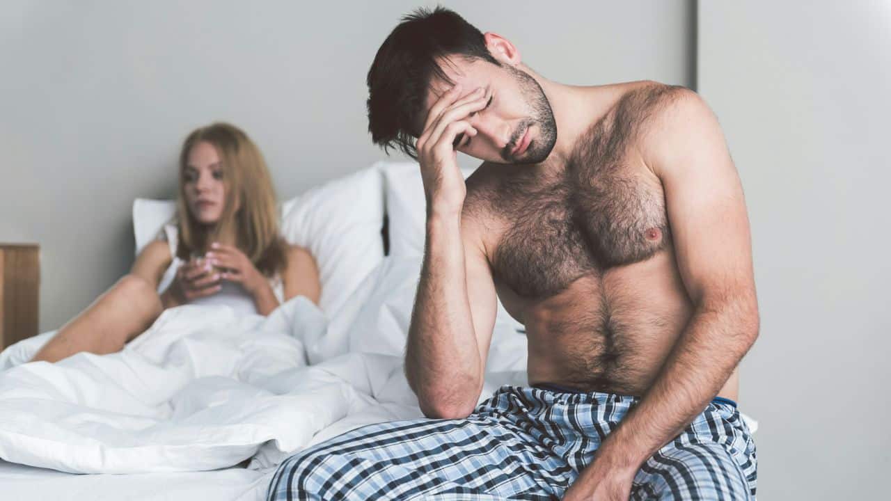 A tired man with his hand on his forehead is sitting on the edge of a bed, while a woman sits in the background.