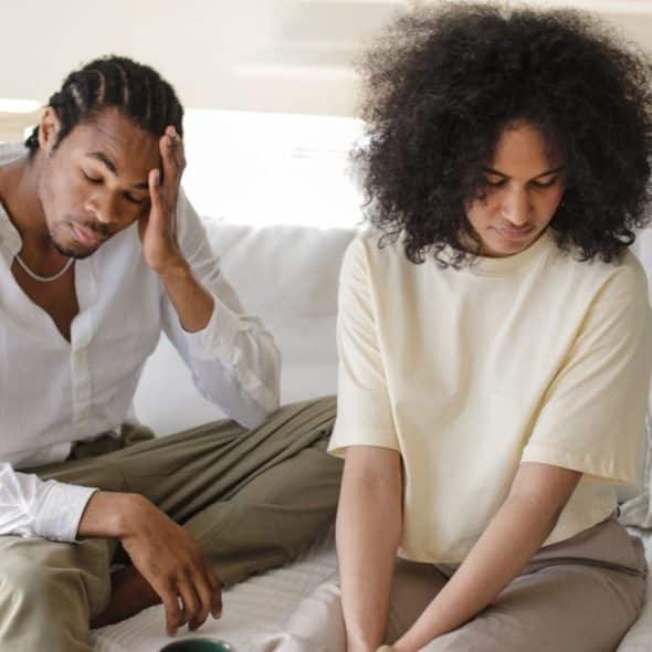 A sad man and woman sit on a couch with their heads down.