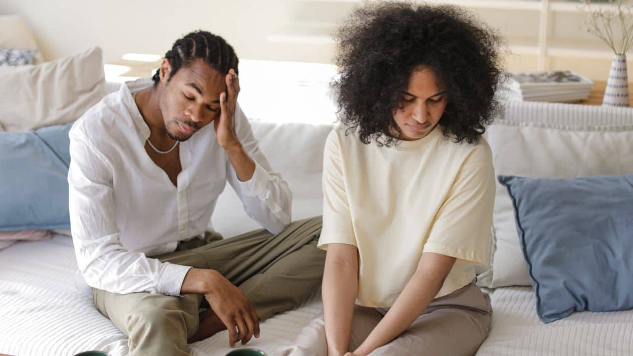 A sad man and woman sit on a couch with their heads down.
