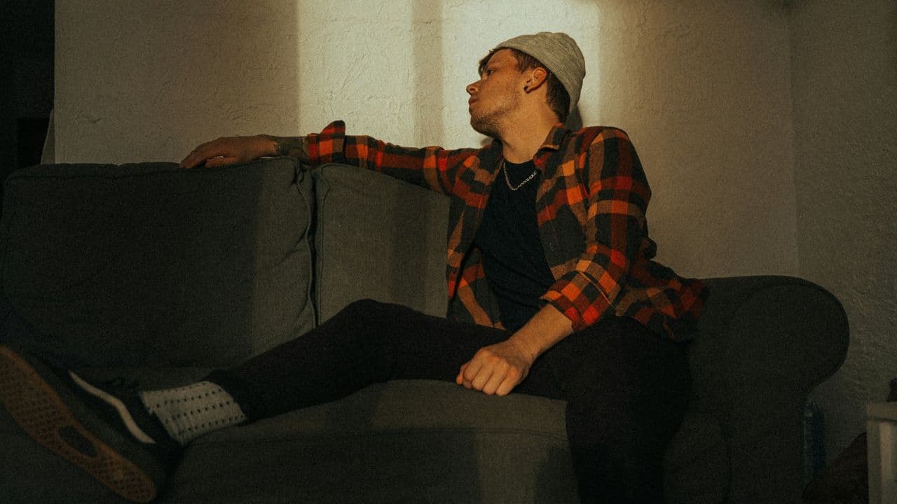 A man in a beanie and plaid shirt sits on a couch and looks to the side.