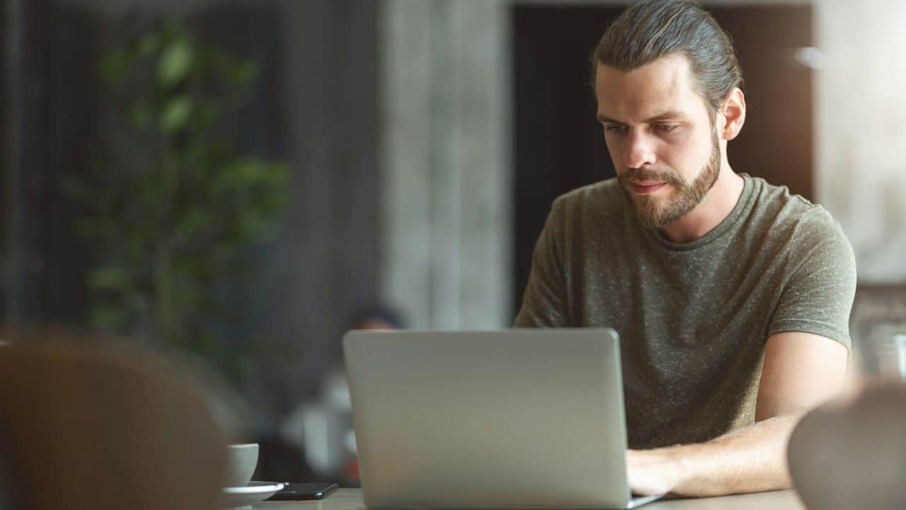 A man with a beard and hair tied back works on a laptop.