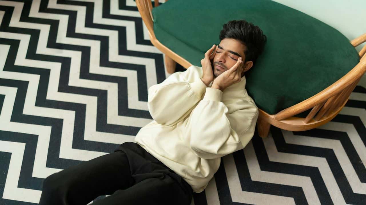 A man in a sweatshirt sits on a zig-zag rug, holding his face in his hands.