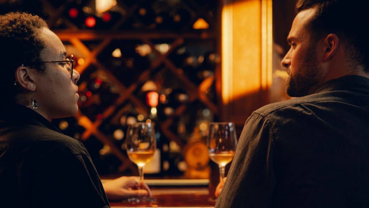 A couple sits across from each other at a table with glasses of white wine.