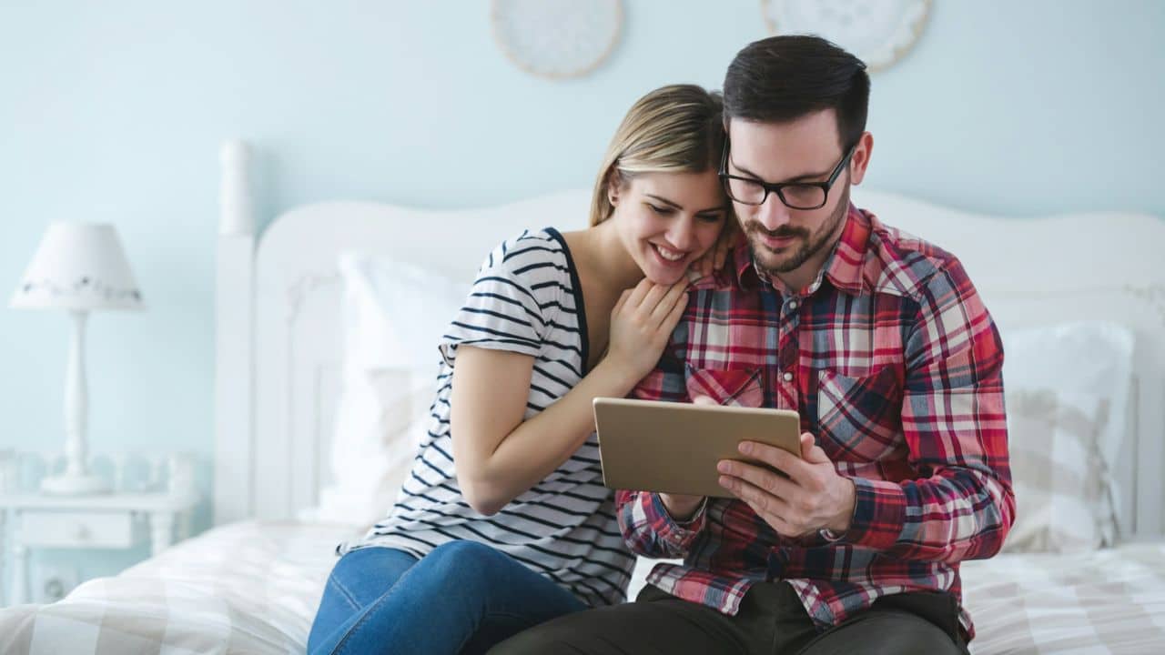 A smiling man and woman sit on a bed and look at a tablet.