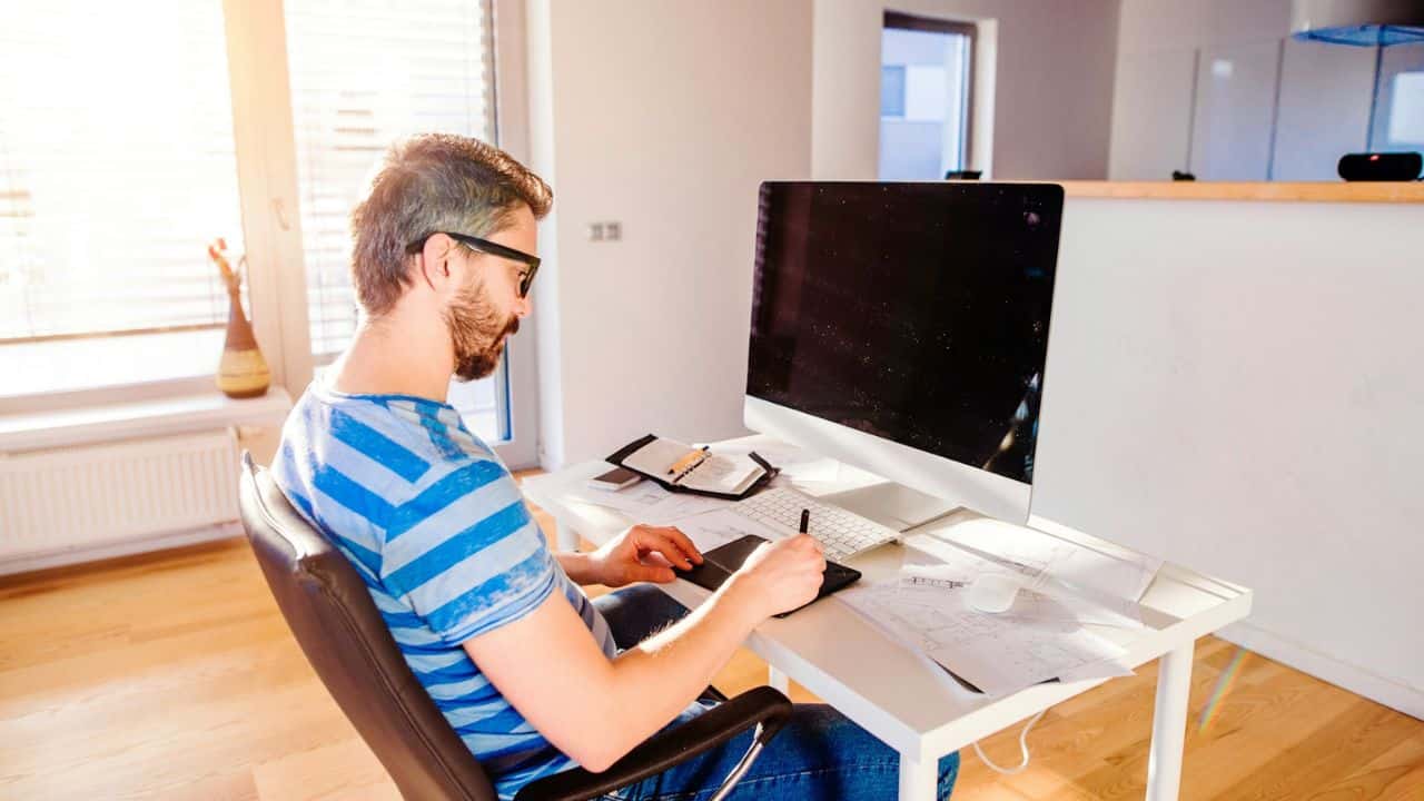 A man with a beard and glasses sits at a desk and works on a digital tablet.