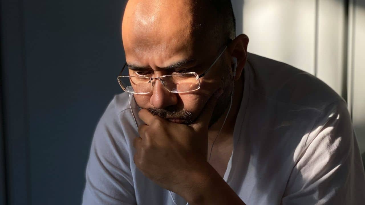 A man with a goatee and glasses sits, looking down, with his hand on his chin.