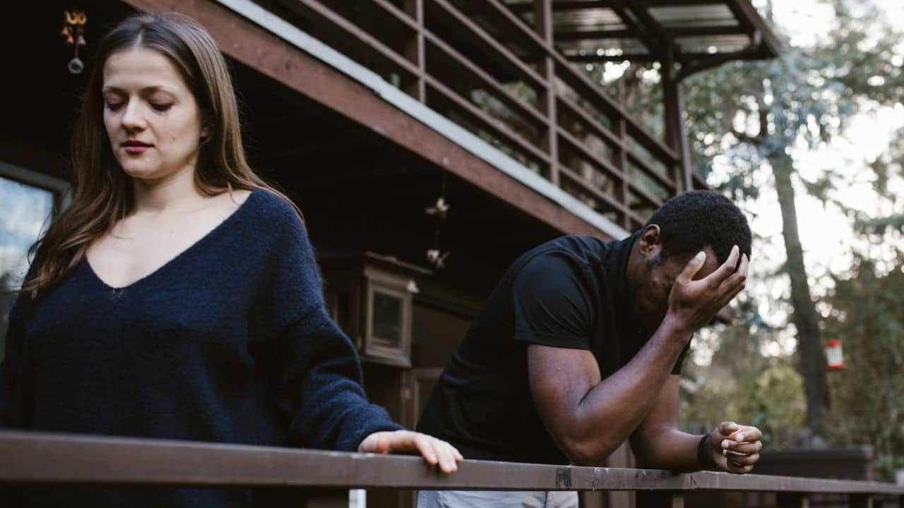 A distressed couple, a woman and a man, stand on a balcony outdoors.