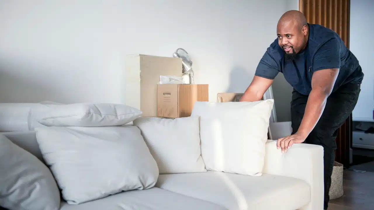 A Black man is pushing a white sofa from behind with a hand on the armrest. There are cardboard boxes behind the sofa.