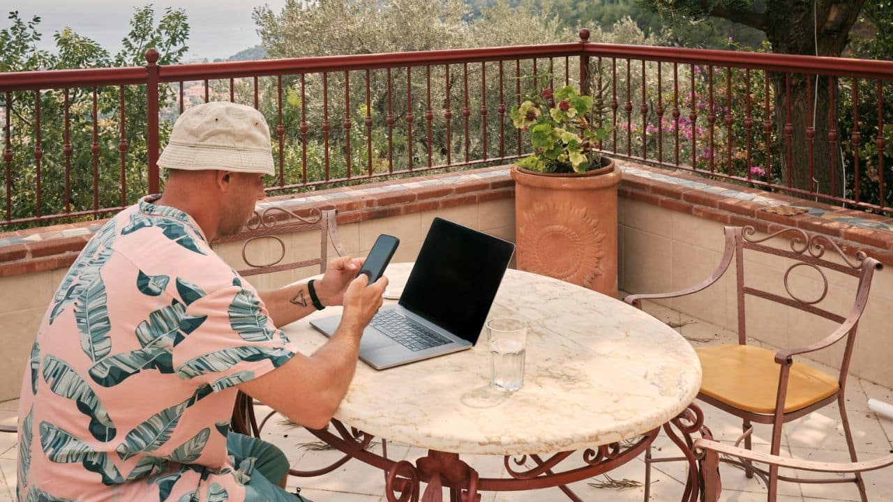 A man on a balcony looks at his phone with a laptop nearby.