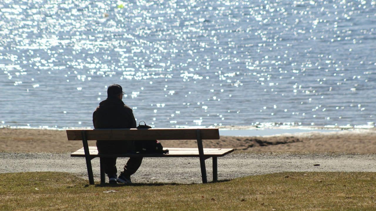 A person wearing a jacket and baseball cap sits on a bench, facing a body of water with sunlight reflecting off the surface.