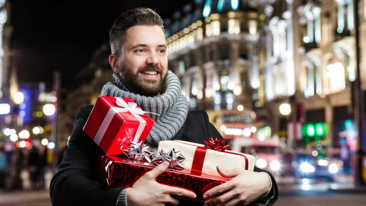 A smiling man with a beard and a gray scarf holds several wrapped gifts while standing on a city street at night.