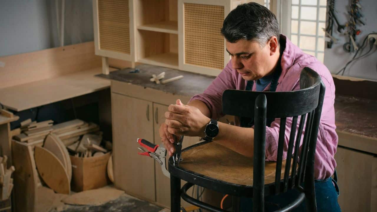 A man is sitting and using pliers to repair a wooden chair in a workshop.