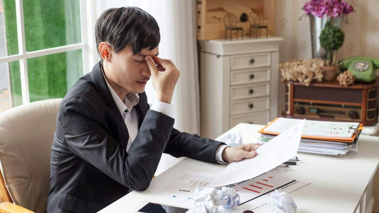 A man in a suit jacket is sitting at a desk and rubbing his eyes with his hand while looking at papers.
