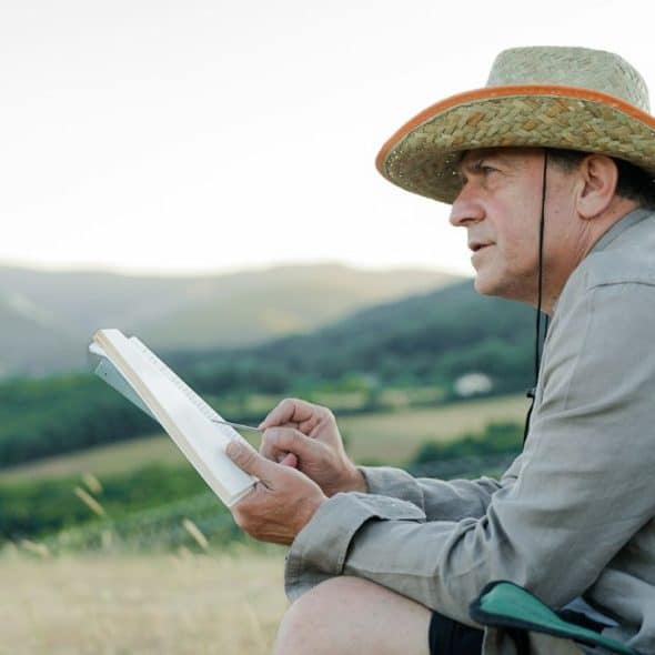 A senior man wearing a straw hat sits and sketches in nature.