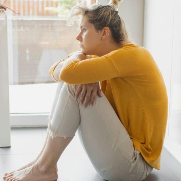 A woman sits on a kitchen floor, hugging her knees and looking away.