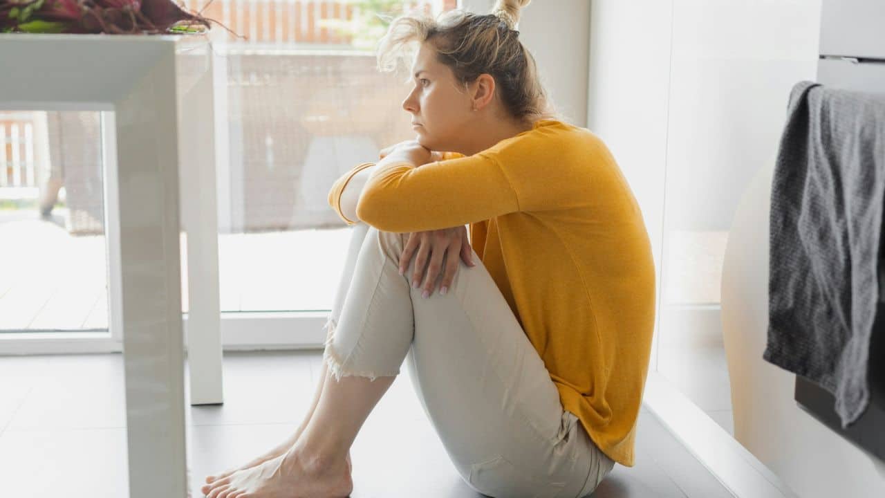 A woman sits on a kitchen floor, hugging her knees and looking away.