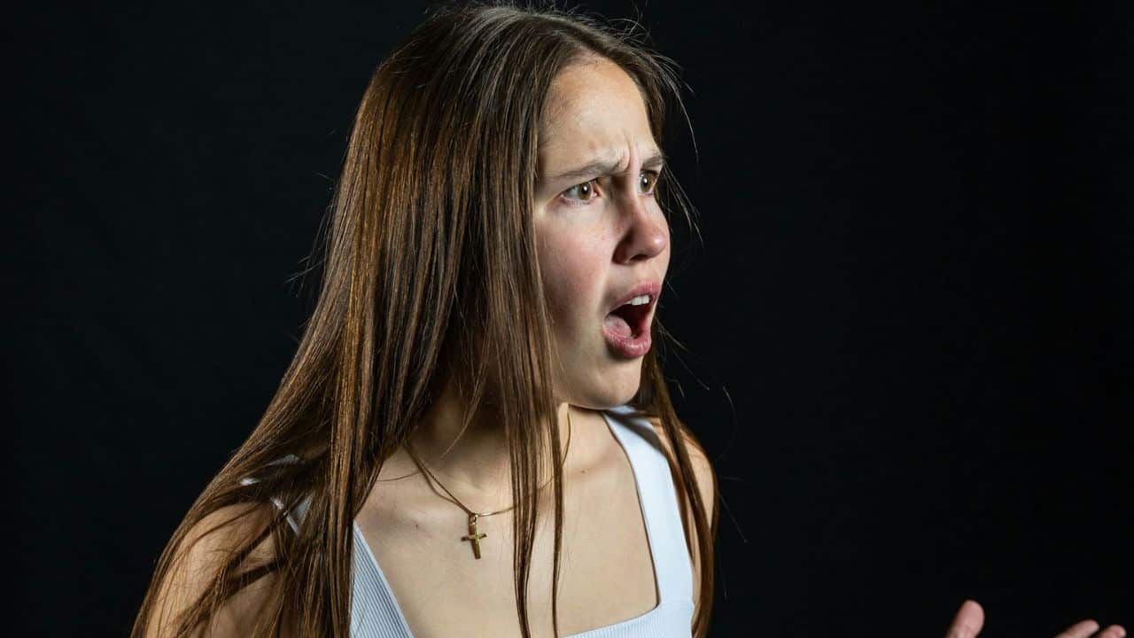 A shocked-looking woman with long brown hair, wearing a white tank top and a cross necklace.