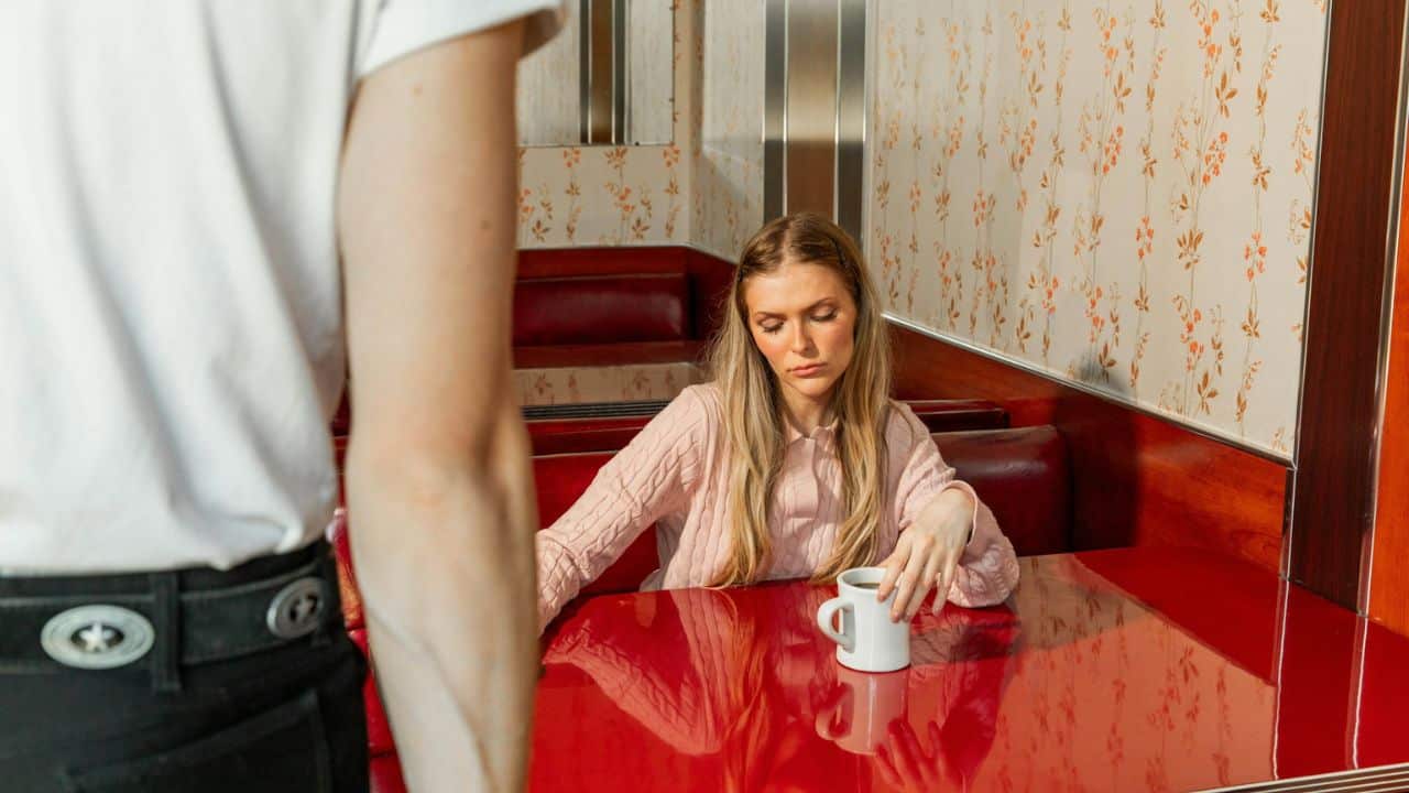 A woman with blond hair sits at a red diner booth, looking down at her mug. The back of a person is visible in the foreground.