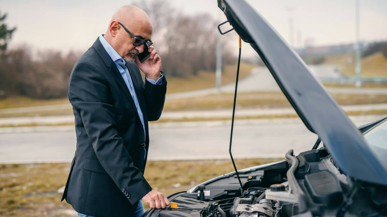 A man in a suit jacket stands with a phone to his ear, looking at an open car hood.