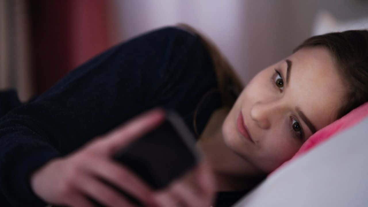 A young woman lying down, holding a phone and looking at the camera.