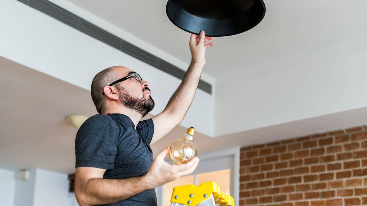 A man on a ladder changing a lightbulb in a black overhead fixture.