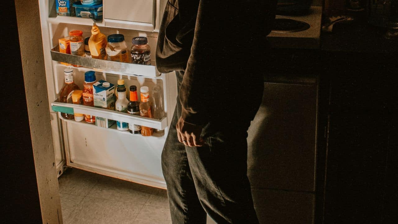 A person's lower body standing next to an open refrigerator full of food.