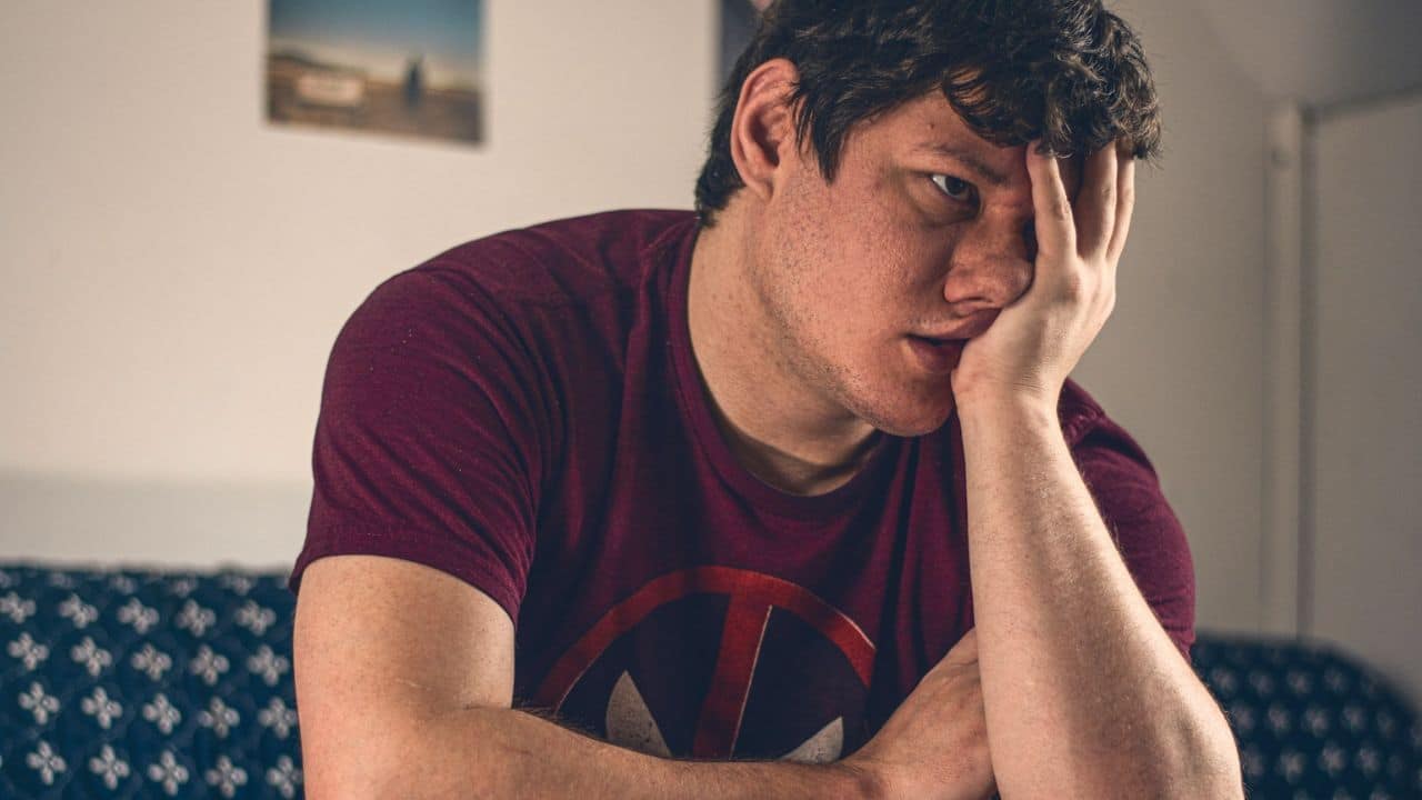 A man in a maroon t-shirt with his hand on his face, looking forward.