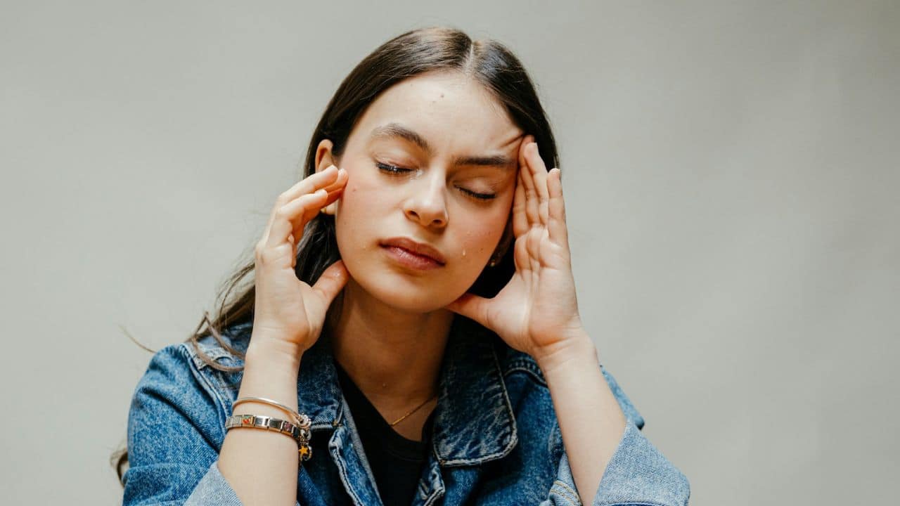A person in a denim jacket with closed eyes and hands on their head.