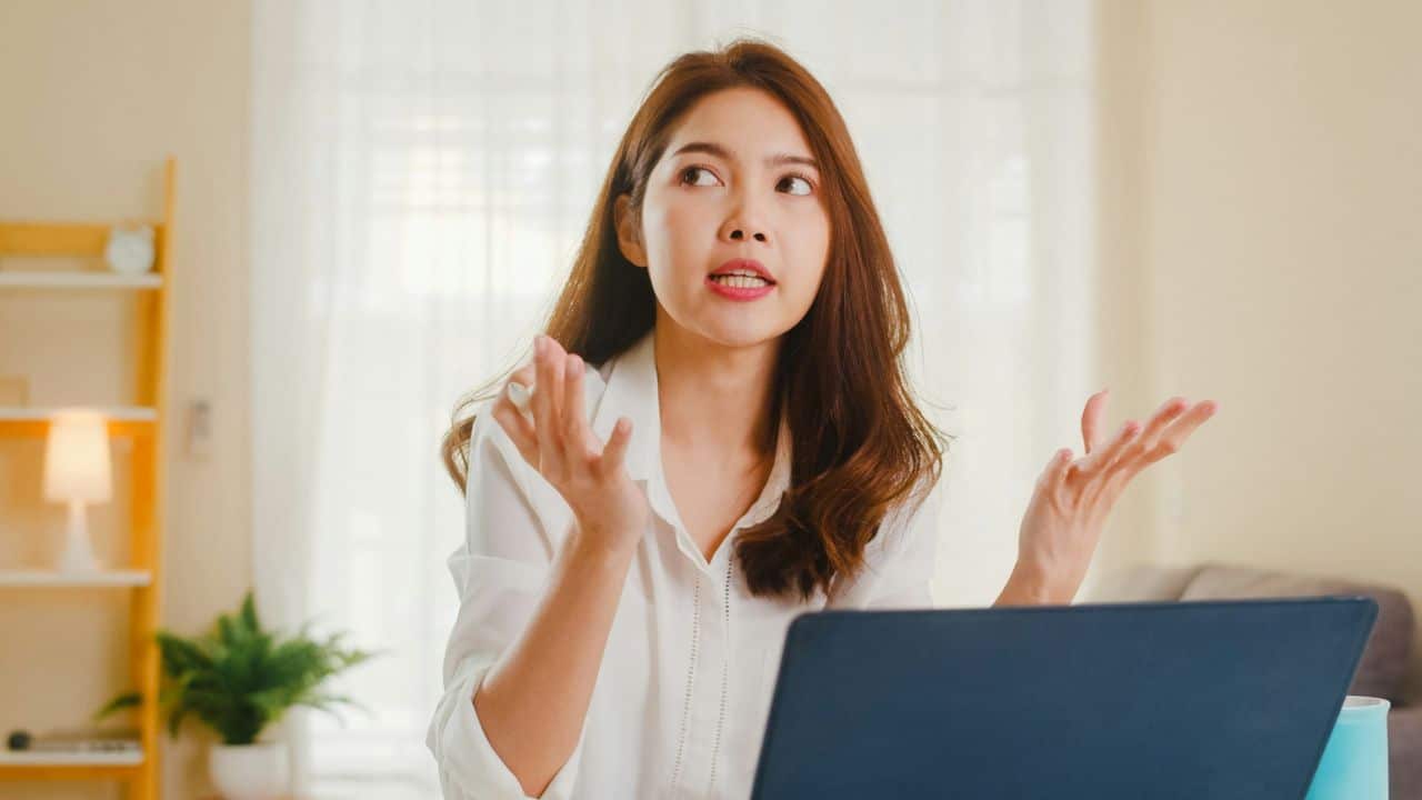 A woman in a white blouse gestures with her hands while seated at a laptop.