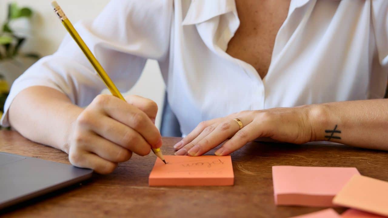 A person's hands writing on orange sticky notes with a yellow pencil.