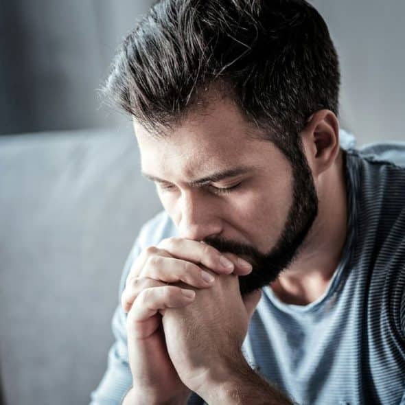 A thoughtful man with a beard sits on a couch with his hands clasped under his chin.